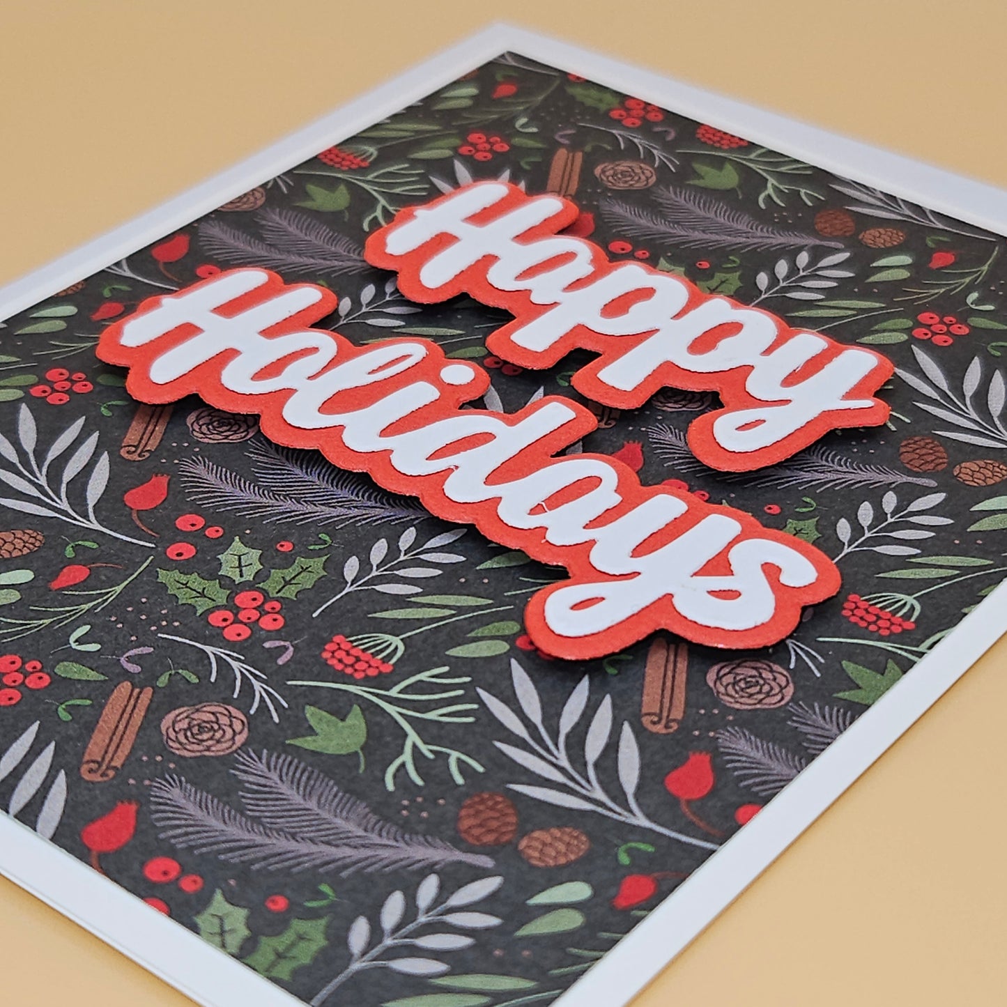A square holiday greeting card with a dark patterned background featuring green leaves, red berries, pinecones, and cinnamon sticks, with large layered 'Happy Holidays' text in red and white. The card is photographed at an angle on a warm beige surface.