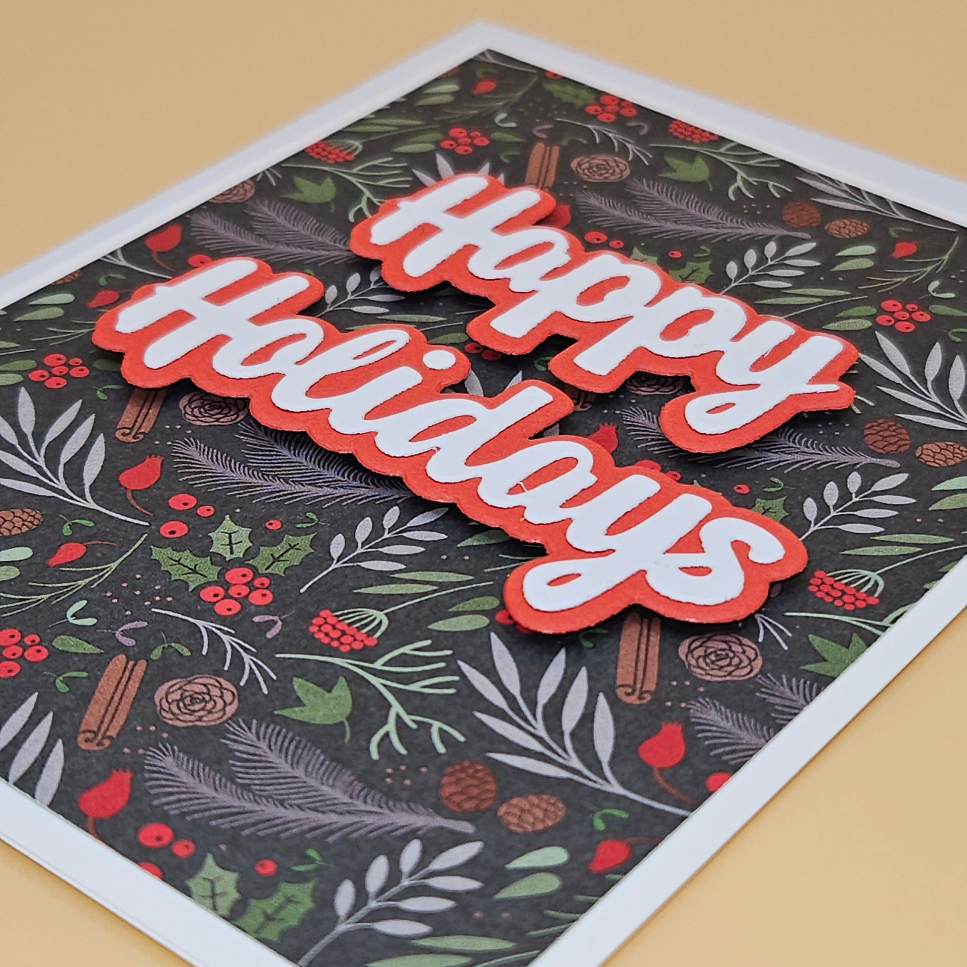 A square holiday greeting card with a dark patterned background featuring green leaves, red berries, pinecones, and cinnamon sticks, with large layered 'Happy Holidays' text in red and white. The card is photographed at an angle on a warm beige surface.
