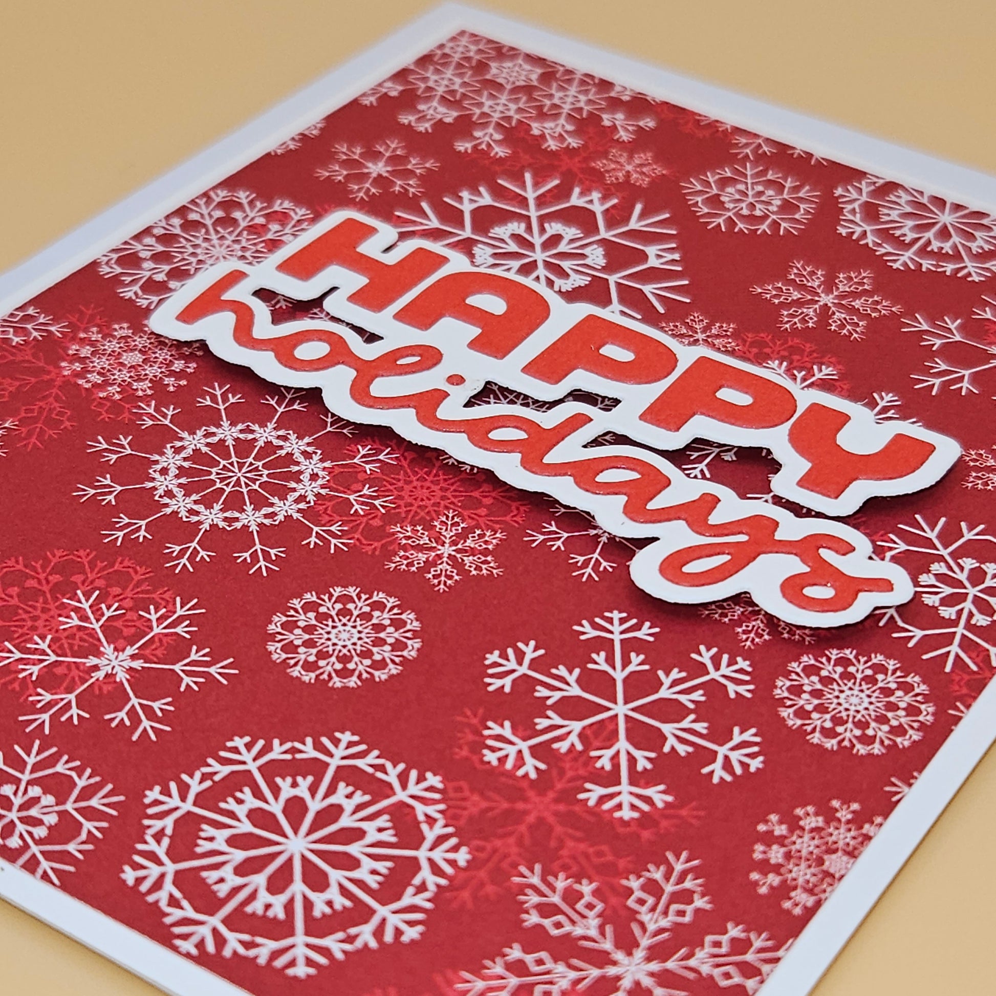 A close-up of a handmade holiday greeting card featuring a red background with white snowflakes and a die-cut 'HAPPY holidays' banner in red and white, placed on a light beige surface.
