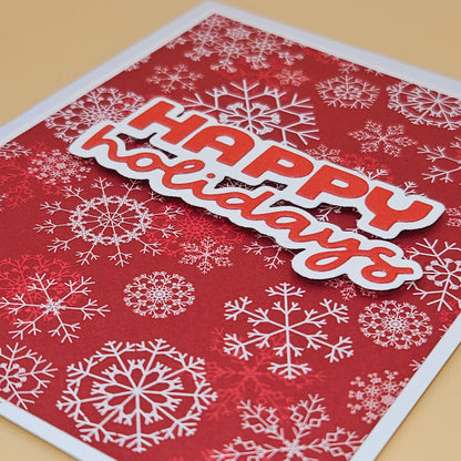 A close-up of a handmade holiday greeting card featuring a red background with white snowflakes and a die-cut 'HAPPY holidays' banner in red and white, placed on a light beige surface.