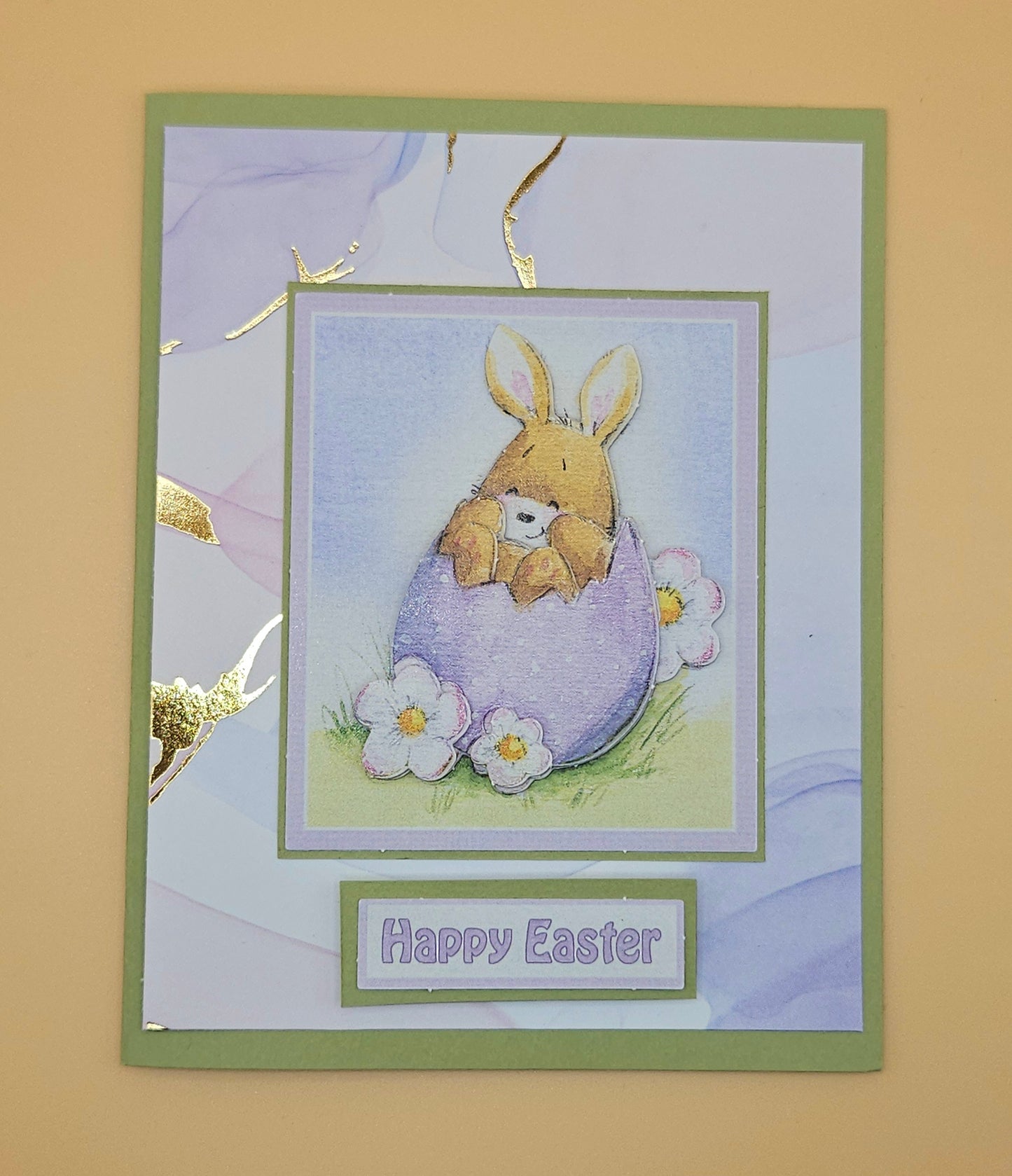 Easter card with a bunny holding a teddy bear in an egg, surrounded by flowers on a beige background.