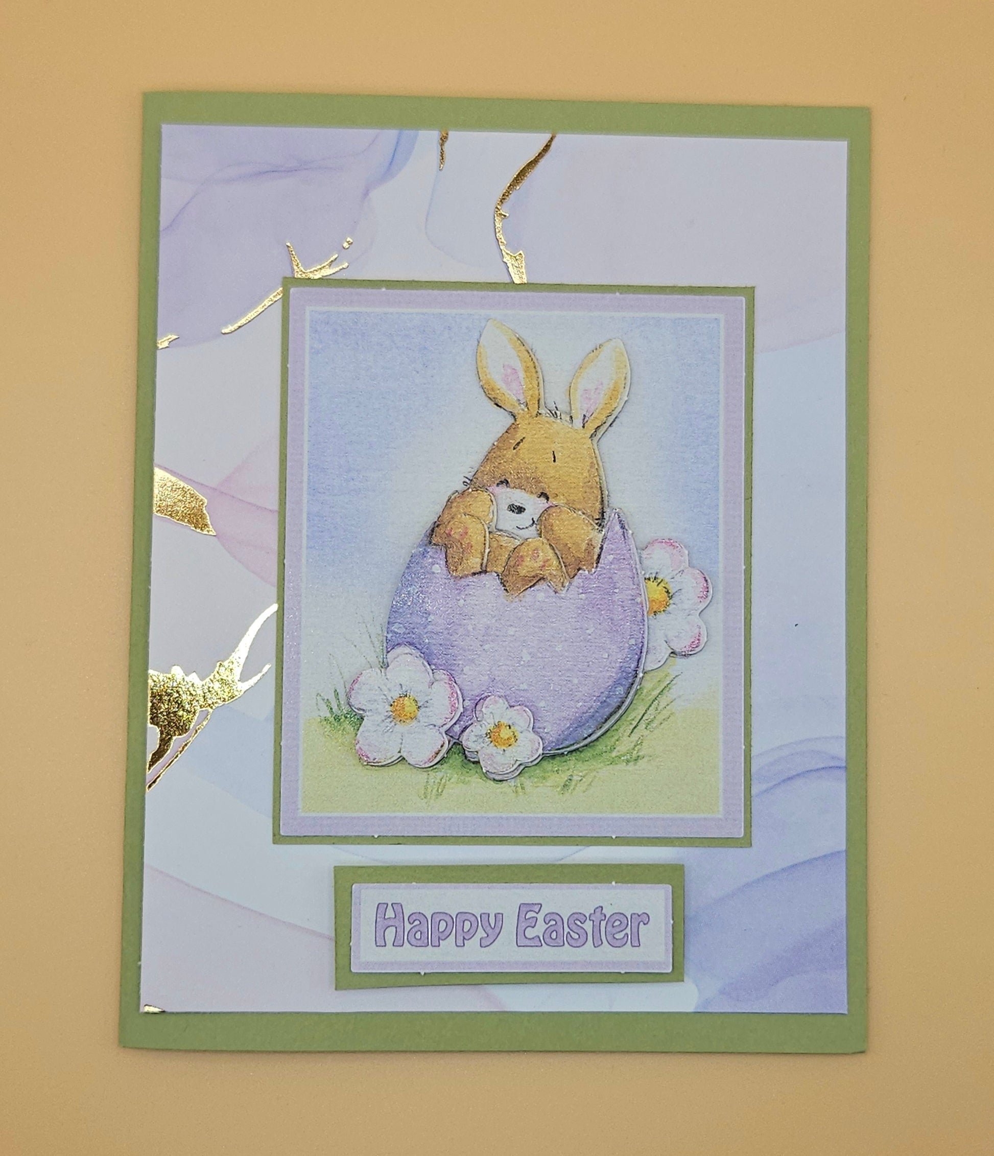 Easter card with a bunny holding a teddy bear in an egg, surrounded by flowers on a beige background.