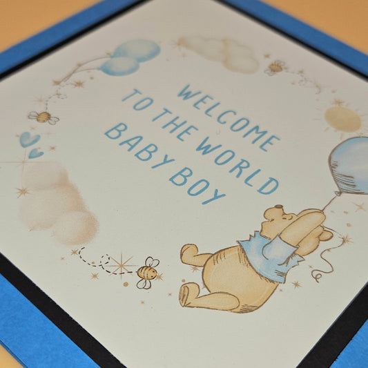 Baby shower card with Winnie the Pooh and text 'Welcome to the world baby boy' on a blue border.