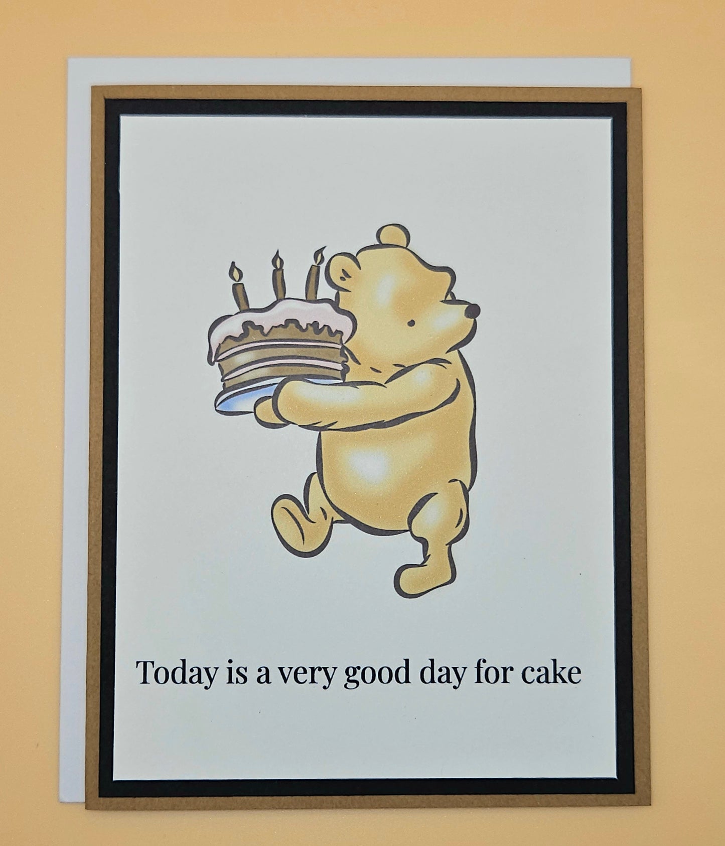 Handmade Winnie the Pooh birthday card with Pooh carrying a birthday cake with lit candles on white inner panel with black and tan border. Text reads Today is a very good day for cake.