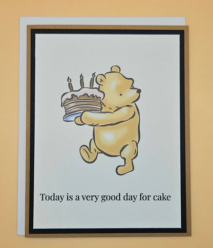 Handmade Winnie the Pooh birthday card with Pooh carrying a birthday cake with lit candles on white inner panel with black and tan border. Text reads Today is a very good day for cake.