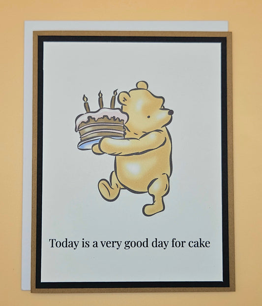 Handmade Winnie the Pooh birthday card with Pooh carrying a birthday cake with lit candles on white inner panel with black and tan border. Text reads Today is a very good day for cake.