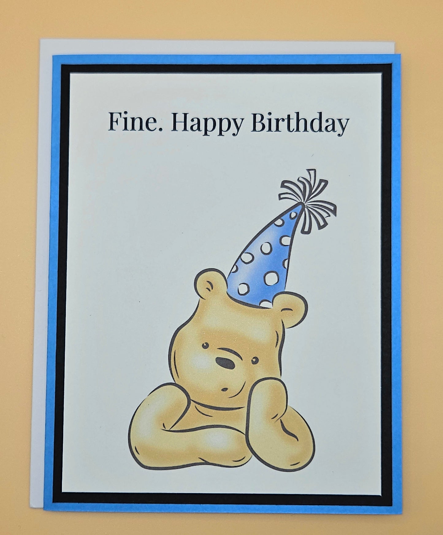 Handmade funny Winnie the Pooh birthday card with grumpy Pooh sitting arms crossed wearing a blue polka dot party hat on white inner panel with black and blue border. Text reads Fine Happy Birthday.
