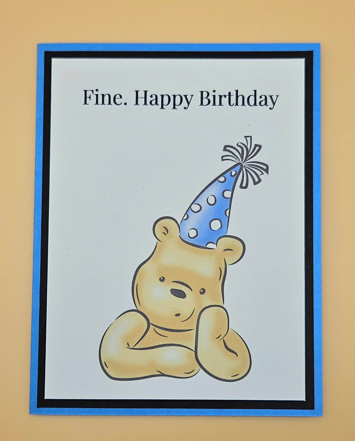 Handmade funny Winnie the Pooh birthday card with grumpy Pooh sitting arms crossed wearing a blue polka dot party hat on white inner panel with black and blue border. Text reads Fine Happy Birthday.