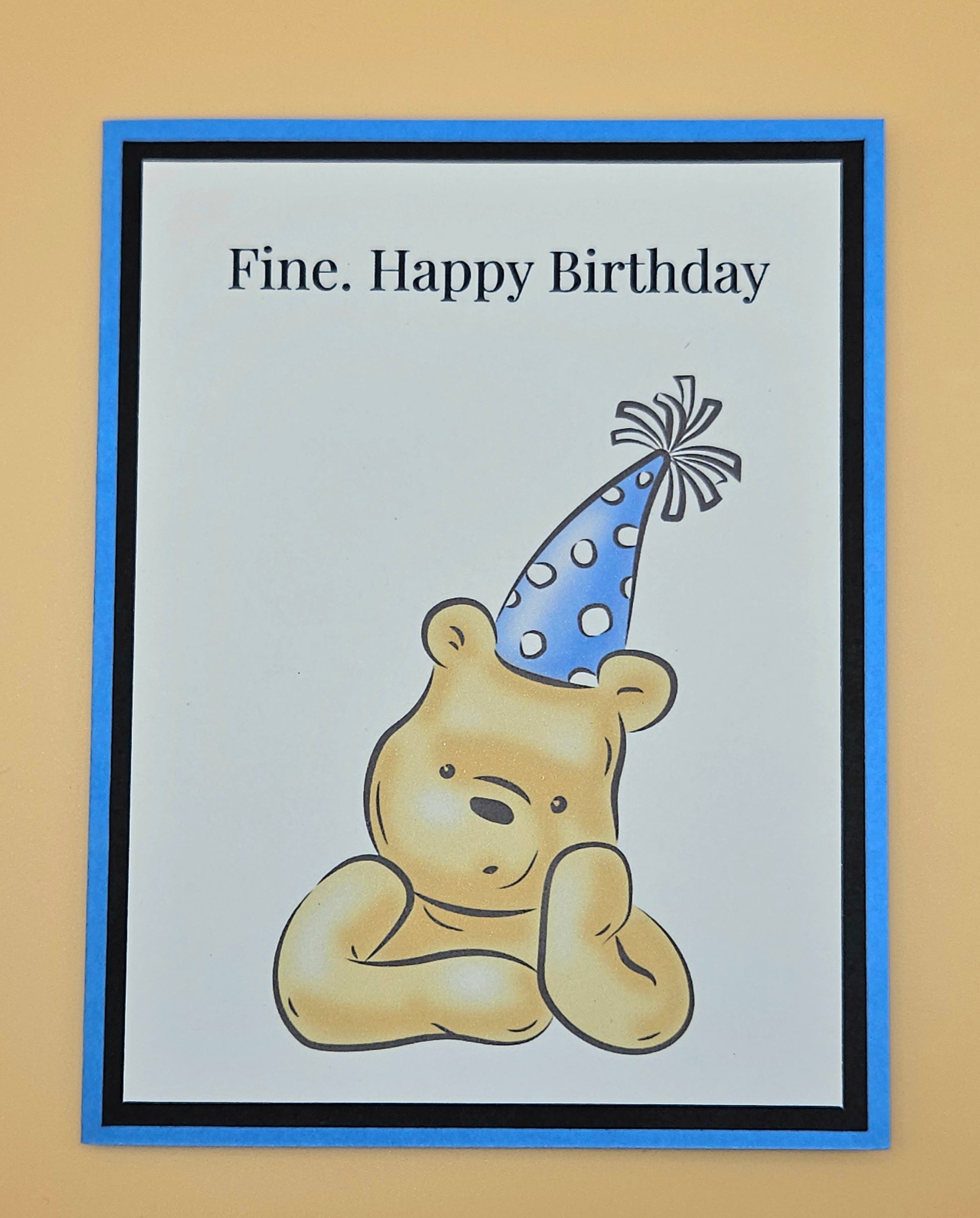 Handmade funny Winnie the Pooh birthday card with grumpy Pooh sitting arms crossed wearing a blue polka dot party hat on white inner panel with black and blue border. Text reads Fine Happy Birthday.