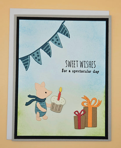 Handmade die-cut Winnie the Pooh birthday card with Piglet holding a cupcake under a bunting banner and gift boxes on light blue and green background. Text reads Sweet Wishes for a spectacular day.