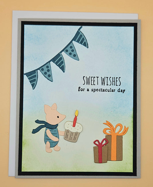 Handmade die-cut Winnie the Pooh birthday card with Piglet holding a cupcake under a bunting banner and gift boxes on light blue and green background. Text reads Sweet Wishes for a spectacular day.