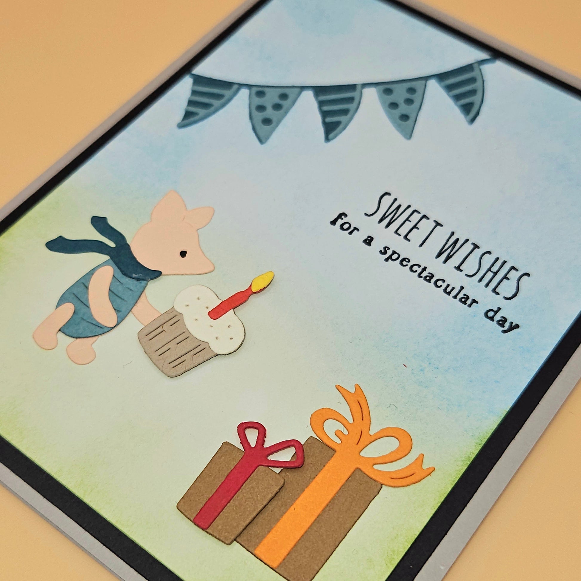 Handmade die-cut Winnie the Pooh birthday card with Piglet holding a cupcake under a bunting banner and gift boxes on light blue and green background. Text reads Sweet Wishes for a spectacular day.