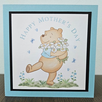 Front of a handmade A2 Mother's Day card featuring a classic storybook sketch illustration of Winnie the Pooh walking while carrying a large bouquet of white daisies and wildflowers, accented with small blue hearts and flying butterflies, finished with a crisp black mat and vibrant sky-blue cardstock frame.