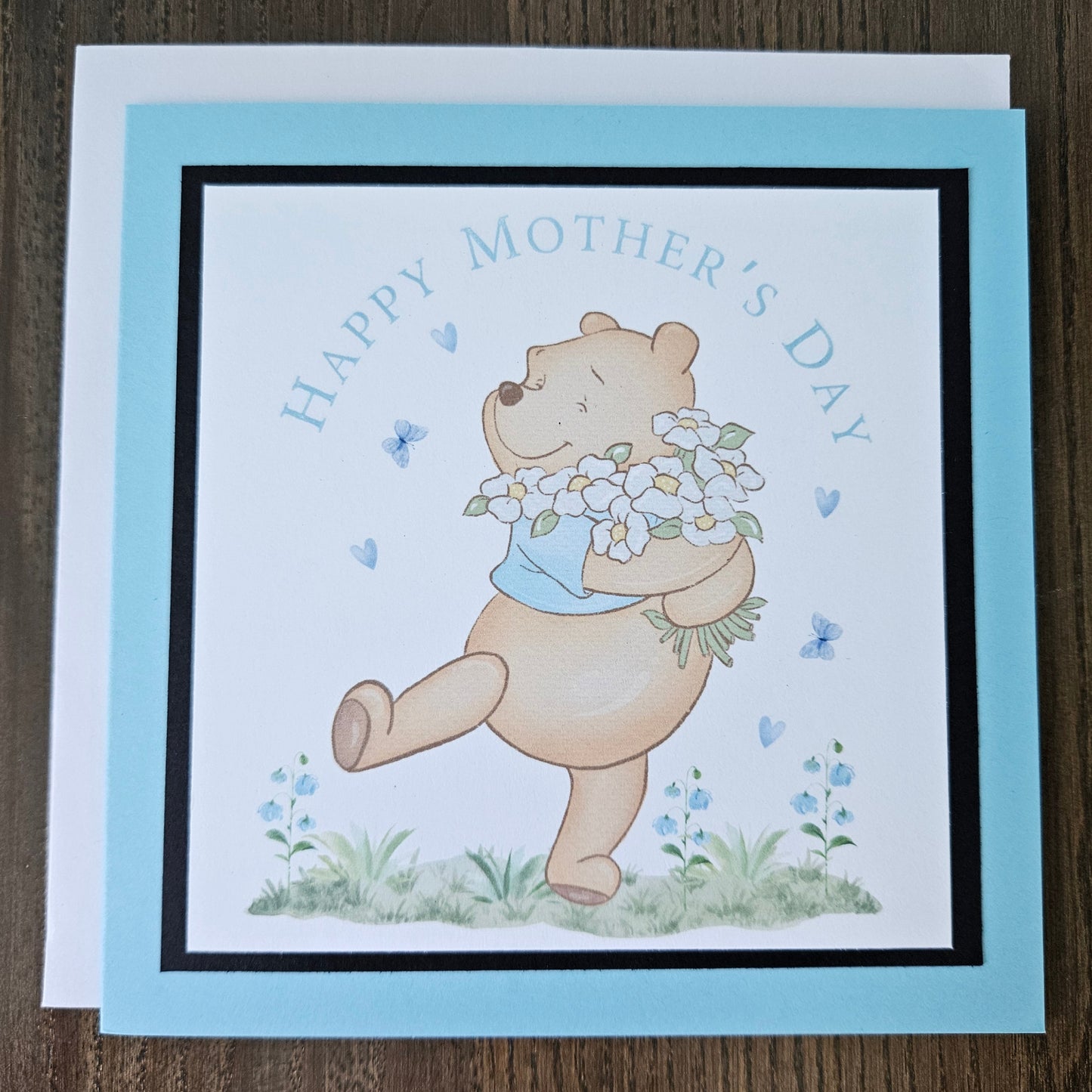 Front of a handmade A2 Mother's Day card featuring a classic storybook sketch illustration of Winnie the Pooh walking while carrying a large bouquet of white daisies and wildflowers, accented with small blue hearts and flying butterflies, finished with a crisp black mat and vibrant sky-blue cardstock frame.