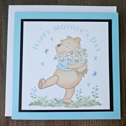 Front of a handmade A2 Mother's Day card featuring a classic storybook sketch illustration of Winnie the Pooh walking while carrying a large bouquet of white daisies and wildflowers, accented with small blue hearts and flying butterflies, finished with a crisp black mat and vibrant sky-blue cardstock frame.