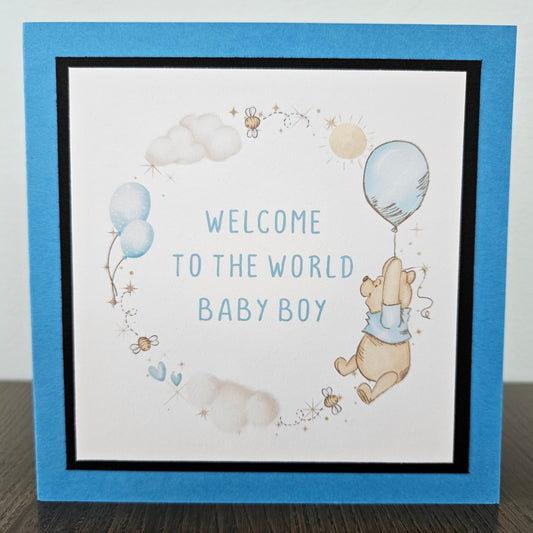 Front of a handmade square baby card featuring a classic Winnie the Pooh illustration floating with a blue balloon inside a circular frame of hand-stenciled clouds, bees, and blue hearts, finished with a black mat and a bright blue card border.