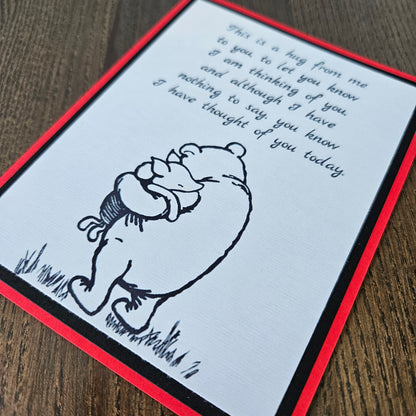 handmade A2 greeting card featuring a classic black ink sketch of Winnie the Pooh hugging Piglet, printed with a touching poem about sending a hug to show someone they are being thought of, framed with a bold black mat and a vibrant red card border.