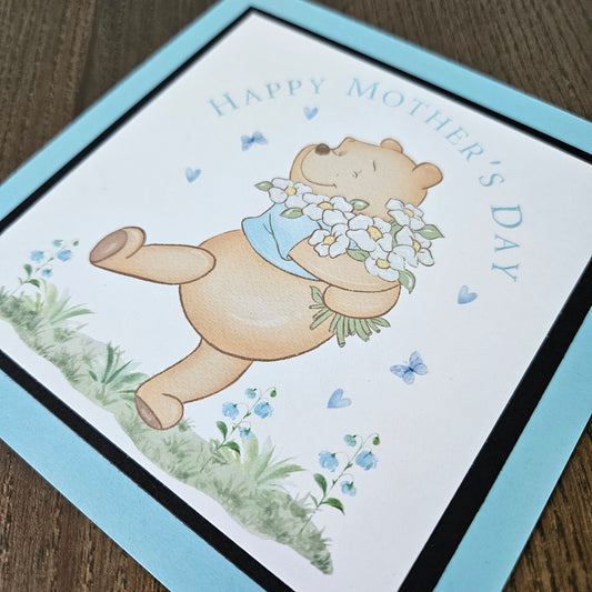 Mother's Day card with a teddy bear holding flowers on a wooden surface