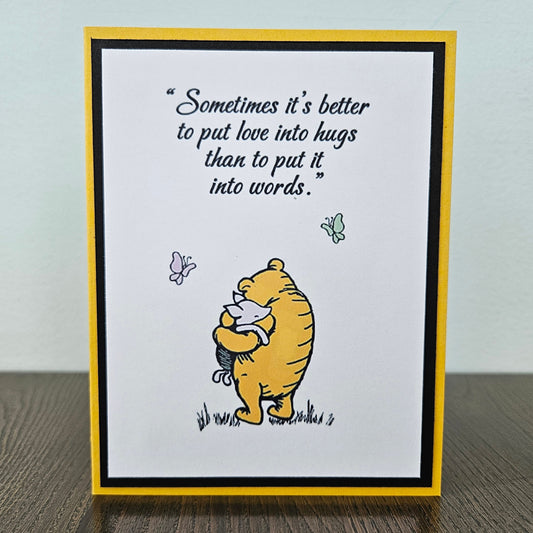 Card with Winnie the Pooh hugging a bear and a quote about love and hugs on a beige background.