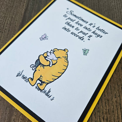 Card with Winnie the Pooh hugging a bear and a quote about love and hugs on a beige background.