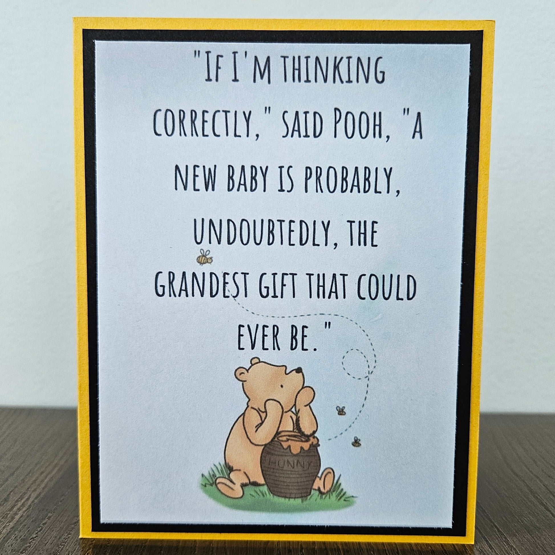 Front of a handmade A2 greeting card featuring a classic Winnie the Pooh sketch illustration sitting with a hunny pot on a soft blue background, showcasing a printed text quote about a new baby being the grandest gift, finished with a crisp black mat frame and a yellow outer card border.