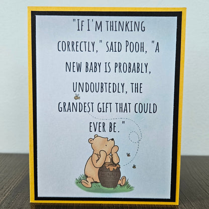 Front of a handmade A2 greeting card featuring a classic Winnie the Pooh sketch illustration sitting with a hunny pot on a soft blue background, showcasing a printed text quote about a new baby being the grandest gift, finished with a crisp black mat frame and a yellow outer card border.