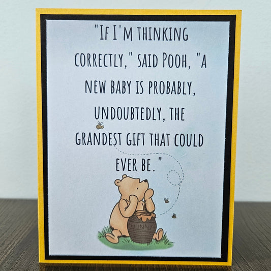 Front of a handmade A2 greeting card featuring a classic Winnie the Pooh sketch illustration sitting with a hunny pot on a soft blue background, showcasing a printed text quote about a new baby being the grandest gift, finished with a crisp black mat frame and a yellow outer card border.