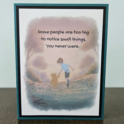 Front of a handmade A2 greeting card featuring a soft watercolor illustration of Winnie the Pooh and Christopher Robin walking together, printed with the quote Some people are too big to notice small things. You never were, finished with a crisp black mat and a rich teal card border.