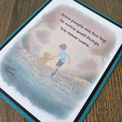 Front of a handmade A2 greeting card featuring a soft watercolor illustration of Winnie the Pooh and Christopher Robin walking together, printed with the quote Some people are too big to notice small things. You never were, finished with a crisp black mat and a rich teal card border.