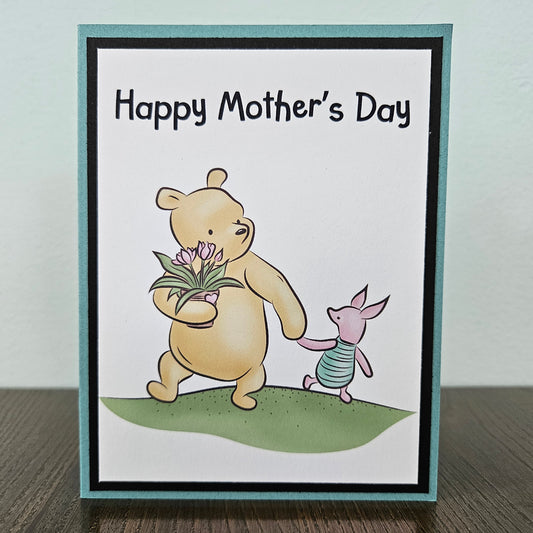 Mother's Day greeting card featuring Winnie the Pooh holding flowers and Piglet walking beside him, with the text 'Happy Mother's Day' at the top. The card has a white center, black border, teal outer border, and a small green ground area.