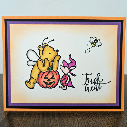 Front of a handmade A2 Halloween card featuring a hand-colored illustration of Winnie the Pooh dressed as a bumblebee and Piglet wearing a purple witch hat next to a jack-o'-lantern pumpkin, printed with the text Trick or Treat, finished with a bold inner mat and layered cardstock frame.