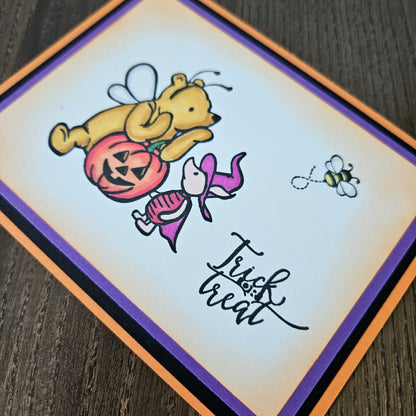 Halloween-themed card with a bear holding a pumpkin and a bee, on a wooden surface.