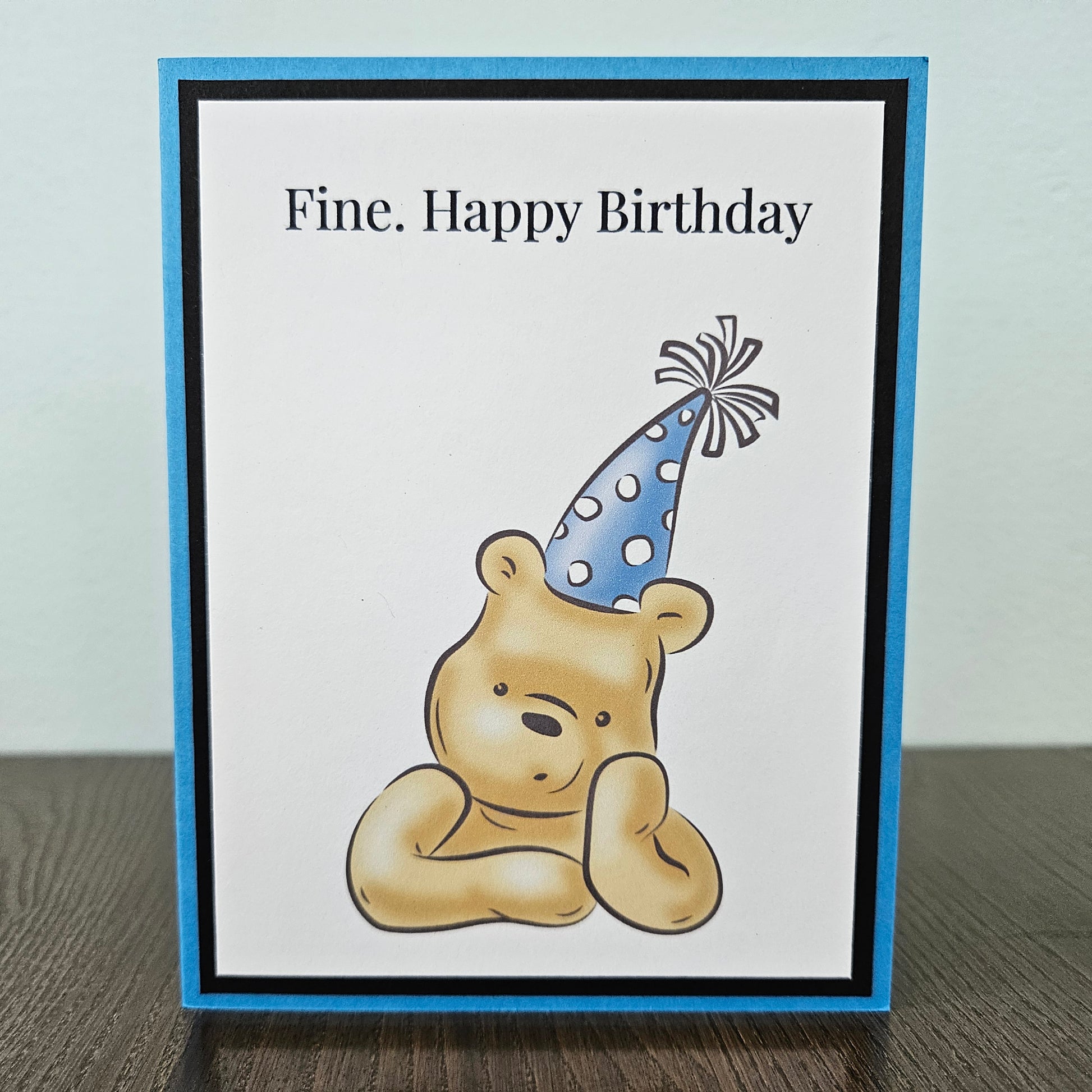 Handmade funny Winnie the Pooh birthday card for him featuring an illustration of a grumpy Pooh Bear wearing a party hat with arms crossed next to a text banner that reads Fine. Happy Birthday, set against a cornflower blue layered border.