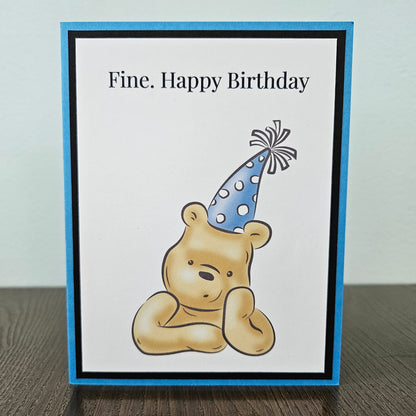 Handmade funny Winnie the Pooh birthday card for him featuring an illustration of a grumpy Pooh Bear wearing a party hat with arms crossed next to a text banner that reads Fine. Happy Birthday, set against a cornflower blue layered border.