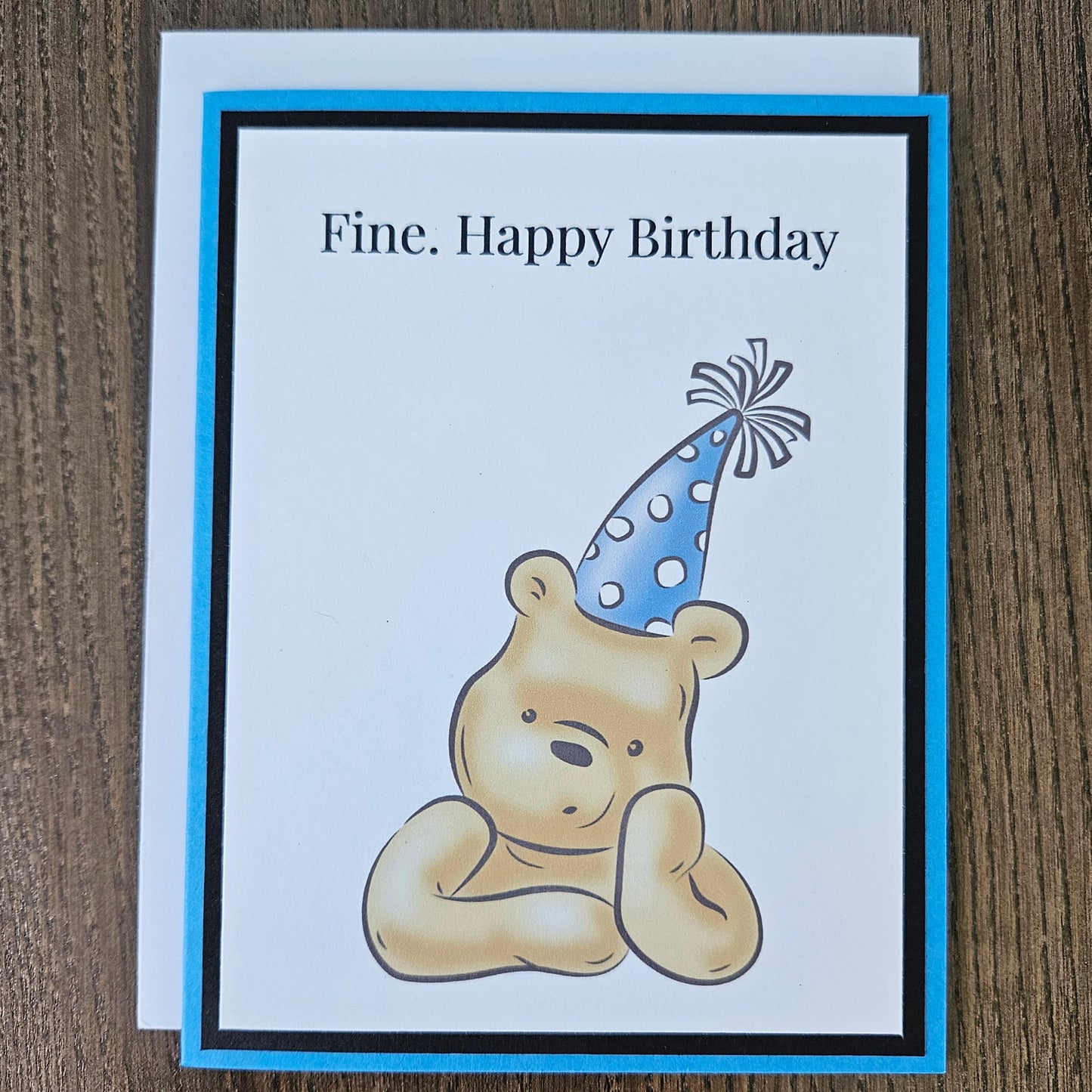 Handmade funny Winnie the Pooh birthday card for him featuring an illustration of a grumpy Pooh Bear wearing a party hat with arms crossed next to a text banner that reads Fine. Happy Birthday, set against a cornflower blue layered border.