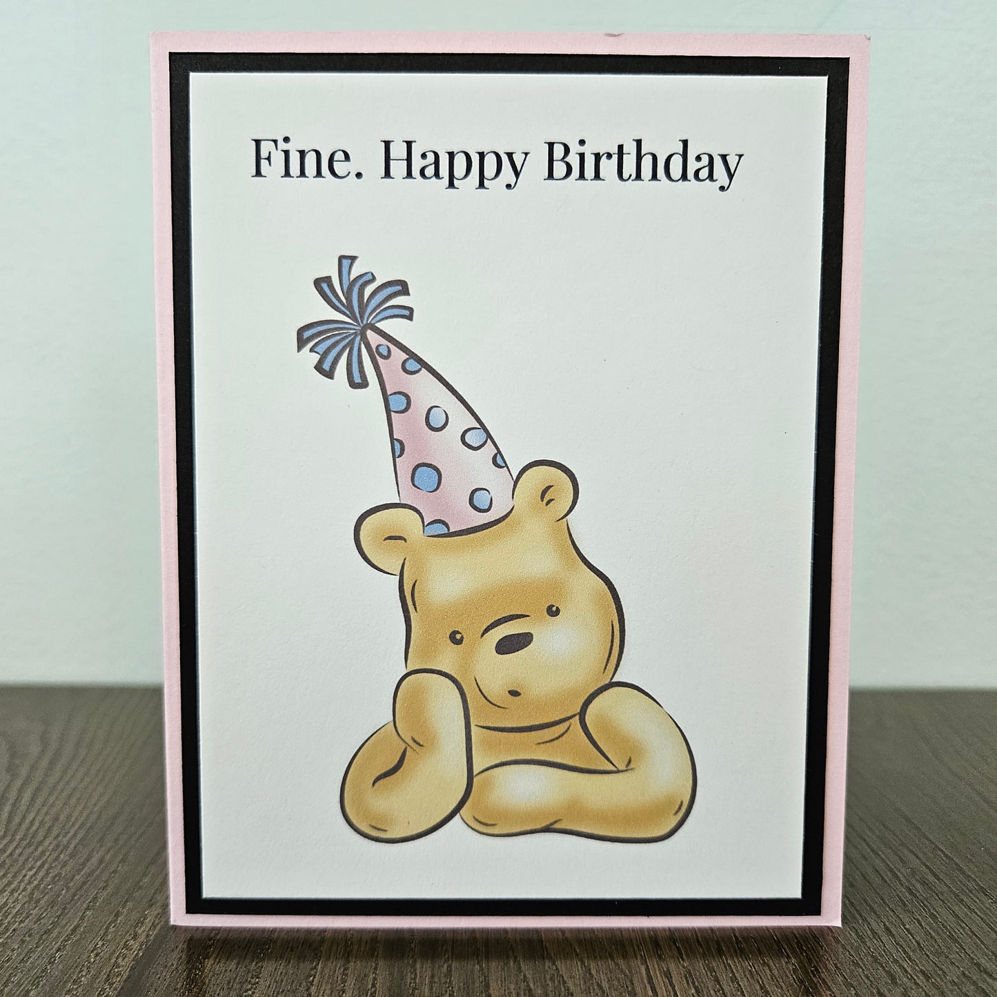 Birthday card with a bear wearing a party hat and text 'Fine. Happy Birthday' on a light background.