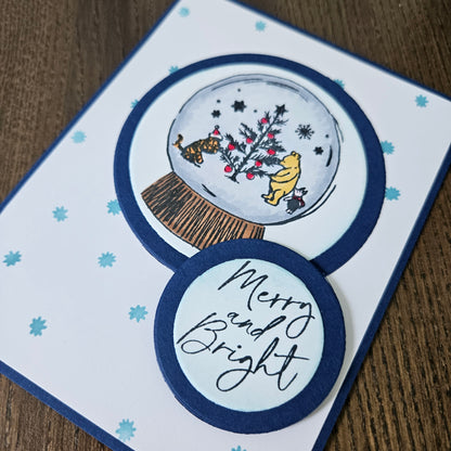 Christmas-themed card with snow globe illustration and 'Merry and Bright' text on a wooden surface.