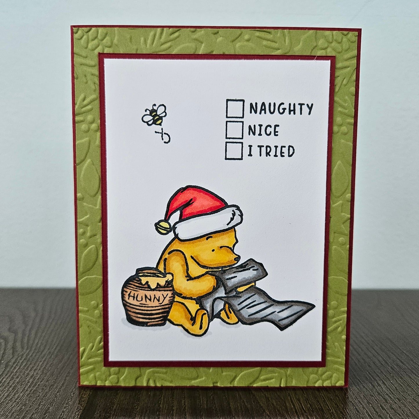 A handmade greeting card featuring Winnie the Pooh wearing a Santa hat and sitting by a honey pot, with the words 'NAUGHTY', 'NICE', and 'I TRIED' next to checkboxes. The card has a white center, a dark red border, and an olive green embossed frame, displayed on a dark wooden surface against a light gray wall.