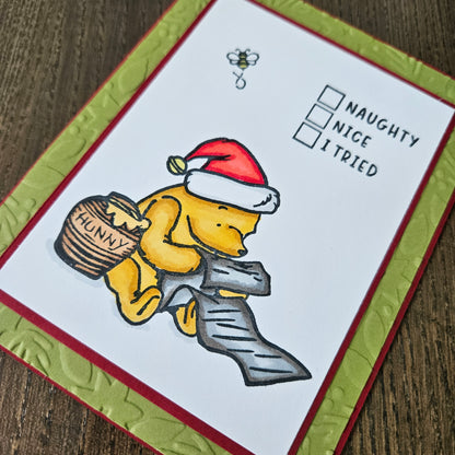 A handmade holiday greeting card featuring Winnie the Pooh holding a basket labeled 'HONEY' while wearing a Santa hat, with the text 'NAUGHTY NICE I TRIED' and a bee illustration. The card has a white center, red border, and olive green embossed border, shown on a wooden surface.