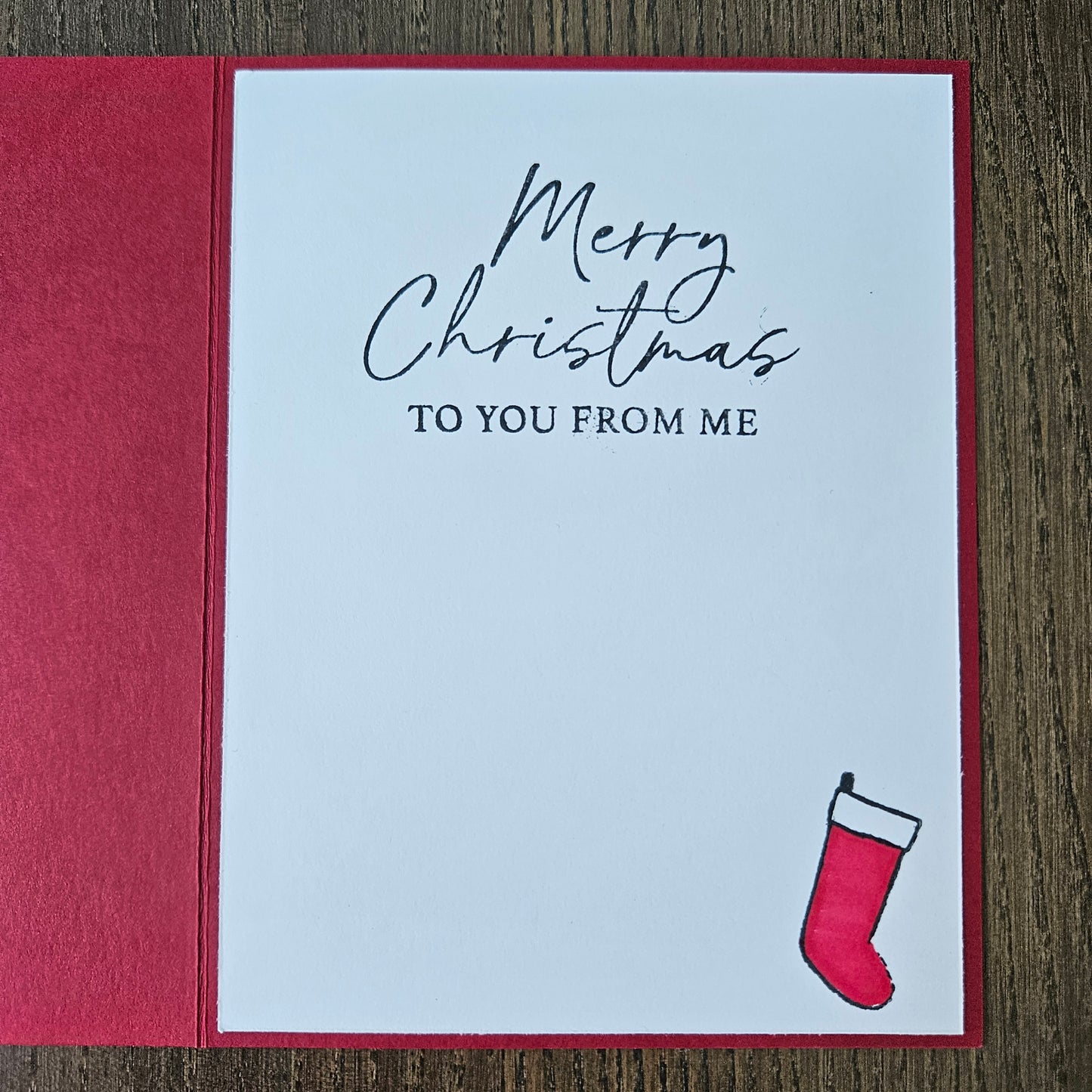 A square Christmas greeting card with a white front and red border, placed on a wooden surface. The card reads 'Merry Christmas TO YOU FROM ME' in black script and serif text, with a small red stocking illustration in the upper right corner.