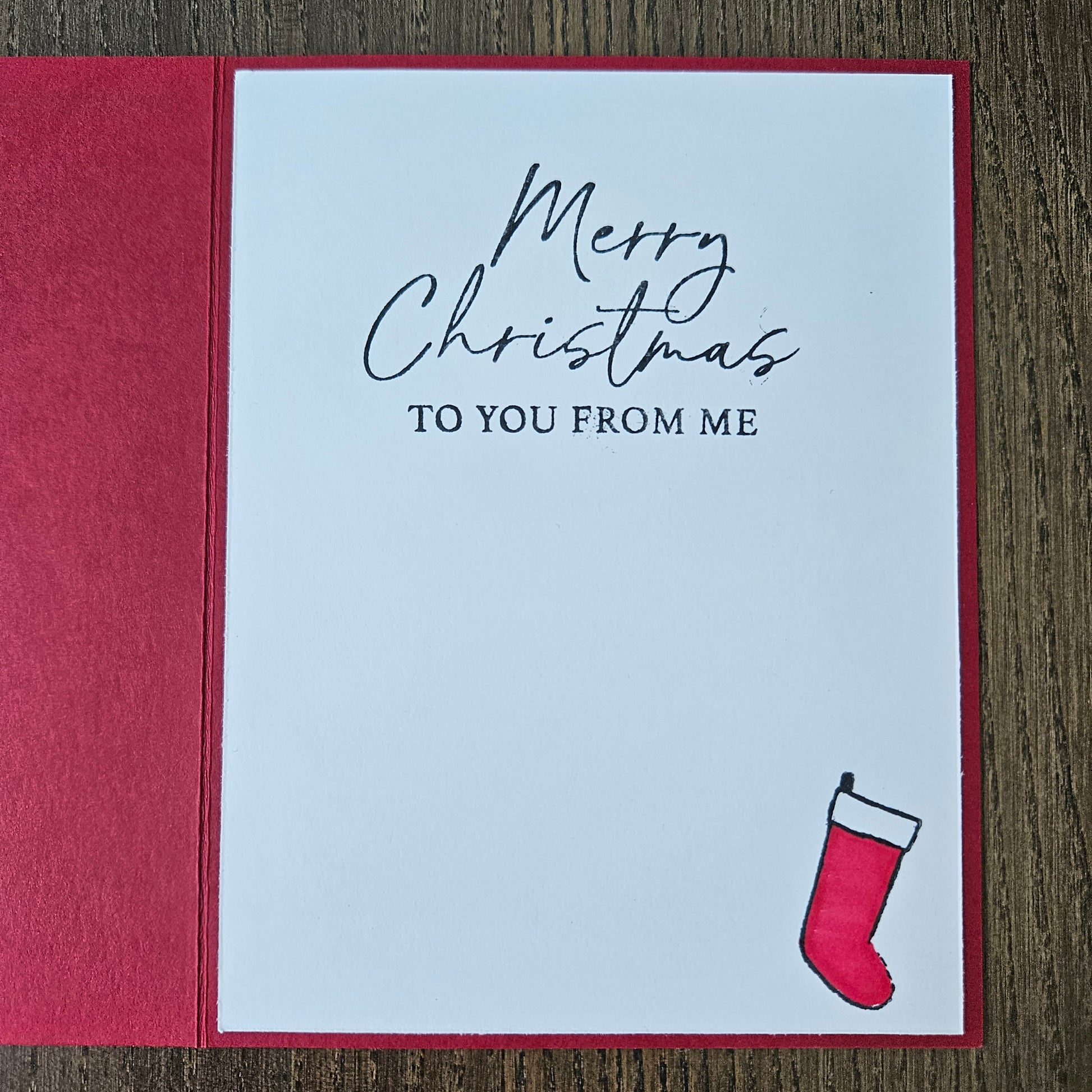 A square Christmas greeting card with a white front and red border, placed on a wooden surface. The card reads 'Merry Christmas TO YOU FROM ME' in black script and serif text, with a small red stocking illustration in the upper right corner.