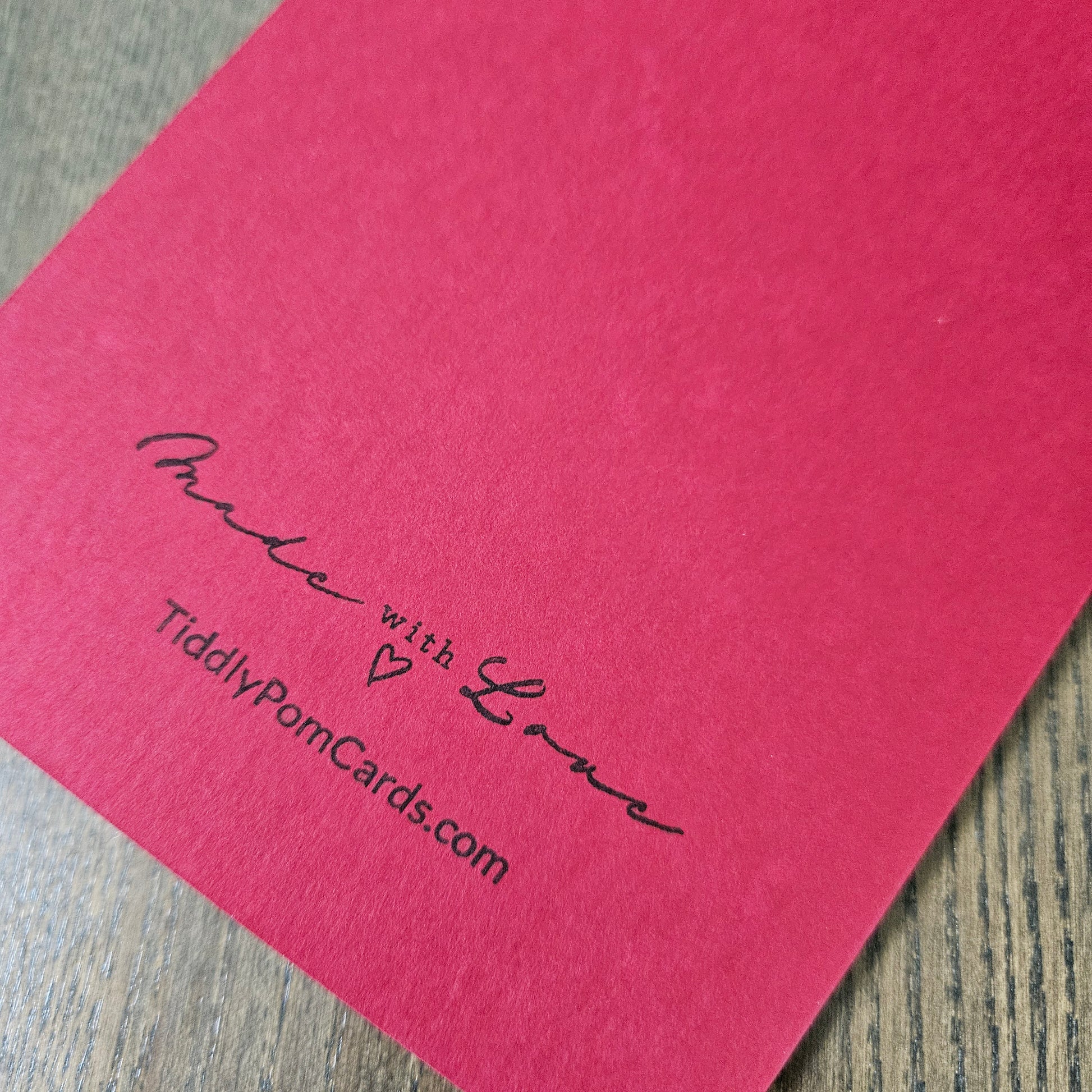A close-up of a textured pink card featuring the printed message 'Made with Love' and the website 'TiddlyPomCards.com' in black ink, resting on a gray wood-grain surface.