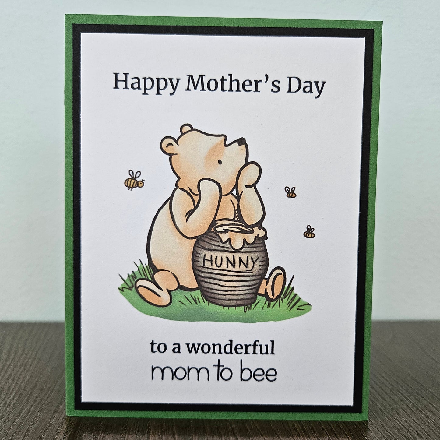 Front of a handmade A2 Mother's Day card featuring a classic storybook sketch illustration of Winnie the Pooh sitting next to a clay honey pot labeled HUNNY on a patch of green grass with three little bees flying around him, printed with the text Happy Mother's Day to a wonderful mom to bee, finished with a bold black inner mat and green cardstock frame