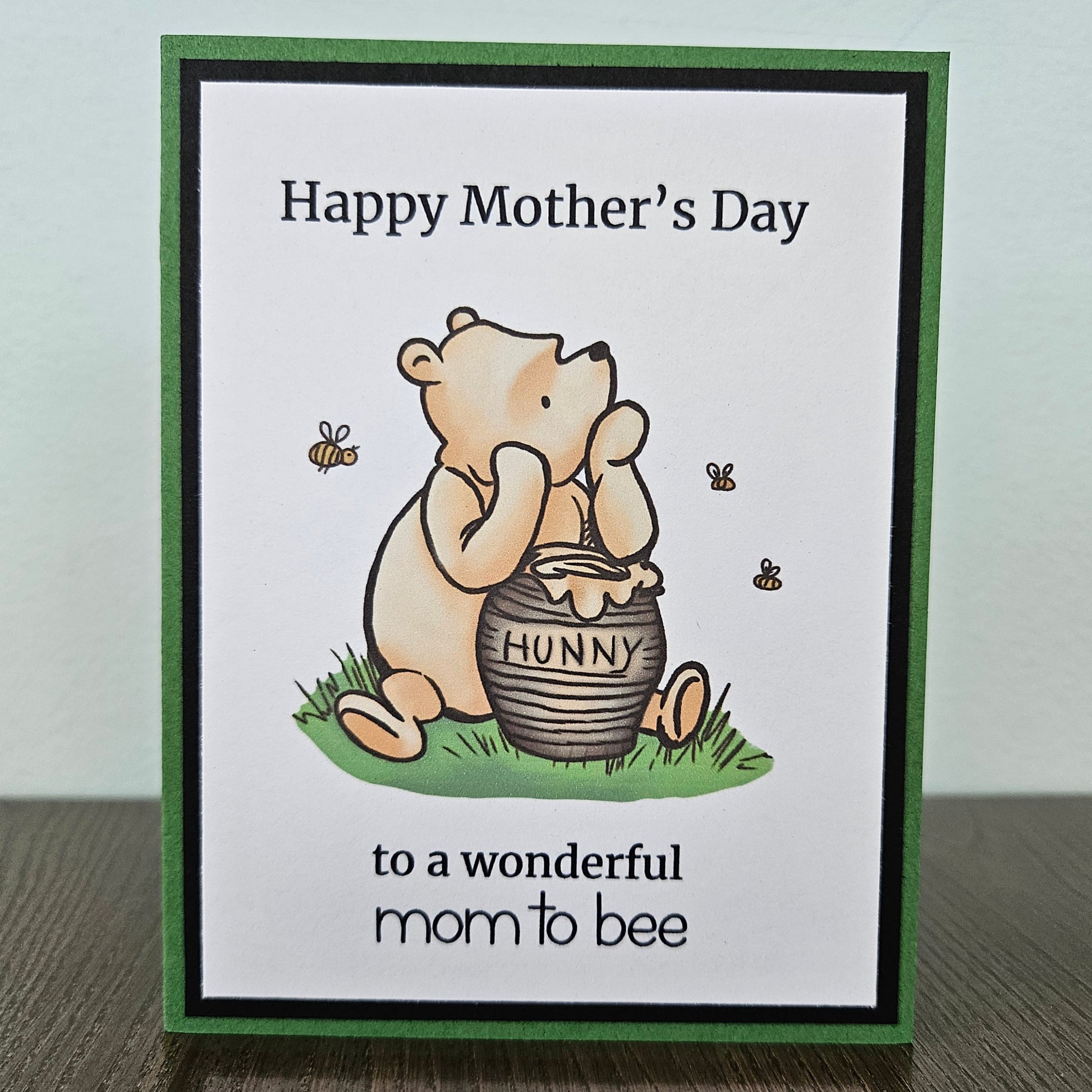 Front of a handmade A2 Mother's Day card featuring a classic storybook sketch illustration of Winnie the Pooh sitting next to a clay honey pot labeled HUNNY on a patch of green grass with three little bees flying around him, printed with the text Happy Mother's Day to a wonderful mom to bee, finished with a bold black inner mat and green cardstock frame