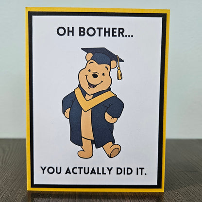 Front of a handmade A2 graduation card featuring a classic storybook sketch illustration of Winnie the Pooh wearing a graduation cap and gown, printed with the funny text Oh Bother... You Actually Did It, finished with a bold black mat and gold cardstock frame.