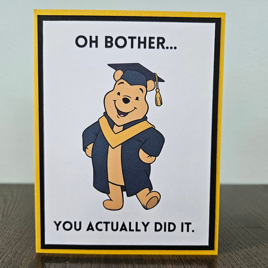 Front of a handmade A2 graduation card featuring a classic storybook sketch illustration of Winnie the Pooh wearing a graduation cap and gown, printed with the funny text Oh Bother... You Actually Did It, finished with a bold black mat and gold cardstock frame.