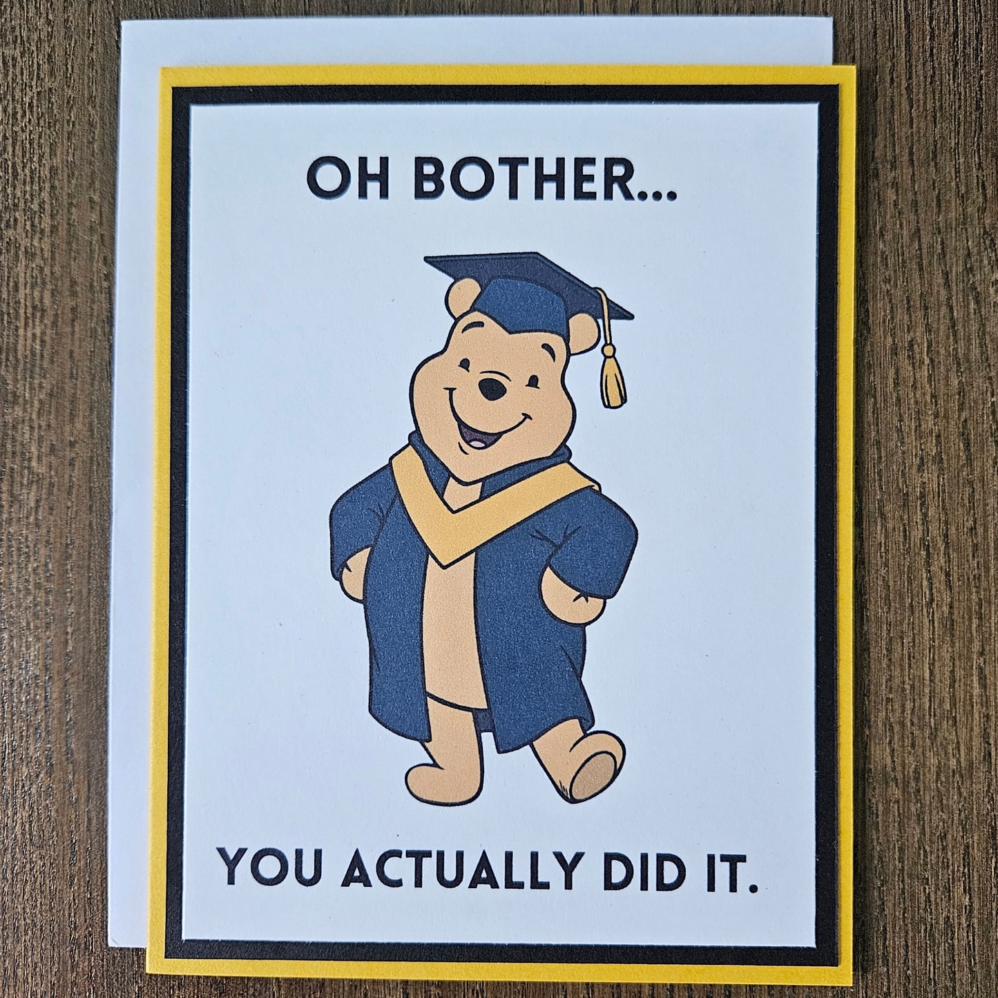 Front of a handmade A2 graduation card featuring a classic storybook sketch illustration of Winnie the Pooh wearing a graduation cap and gown, printed with the funny text Oh Bother... You Actually Did It, finished with a bold black mat and gold cardstock frame.