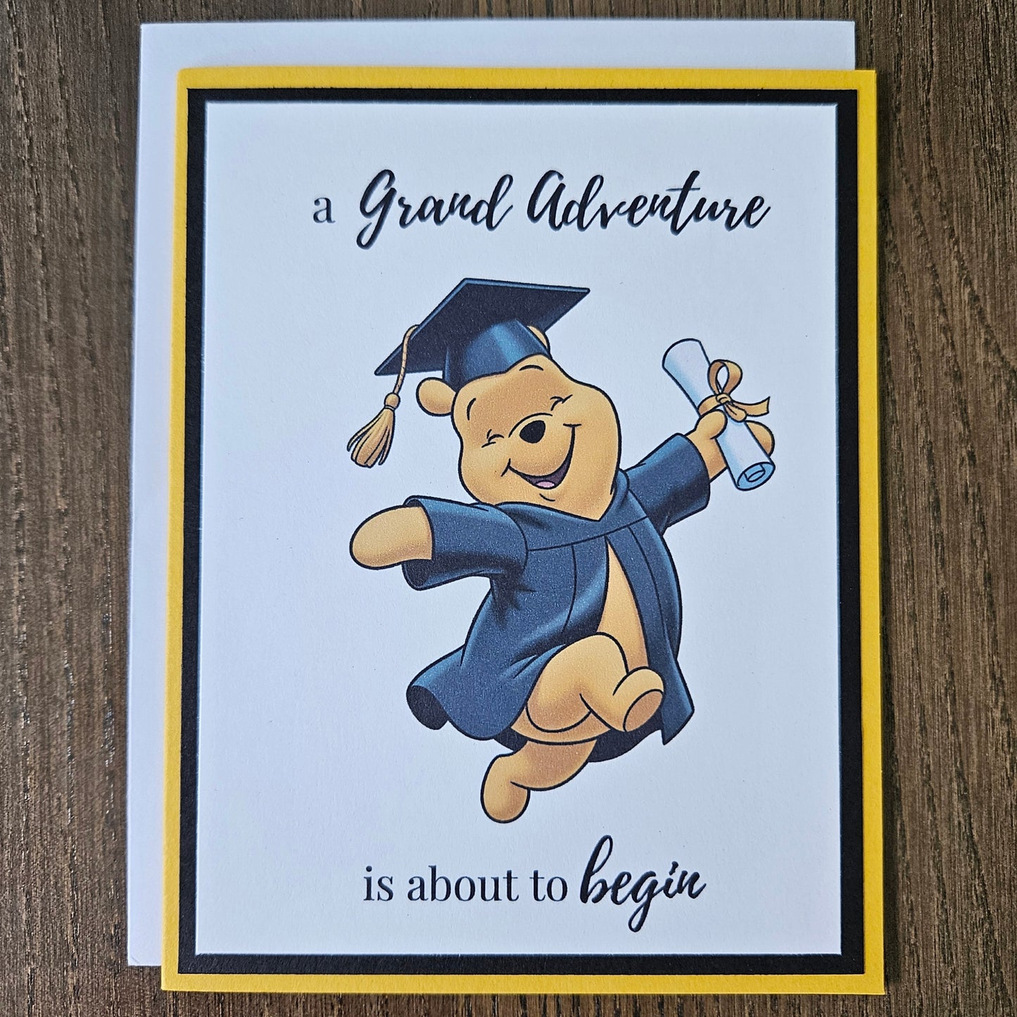 Front of a handmade A2 graduation card featuring a classic storybook sketch illustration of Winnie the Pooh leaping joyfully while wearing a graduation cap and gown and holding a diploma, printed with the text A Grand Adventure is About to Begin, finished with a bold black mat and gold cardstock frame.