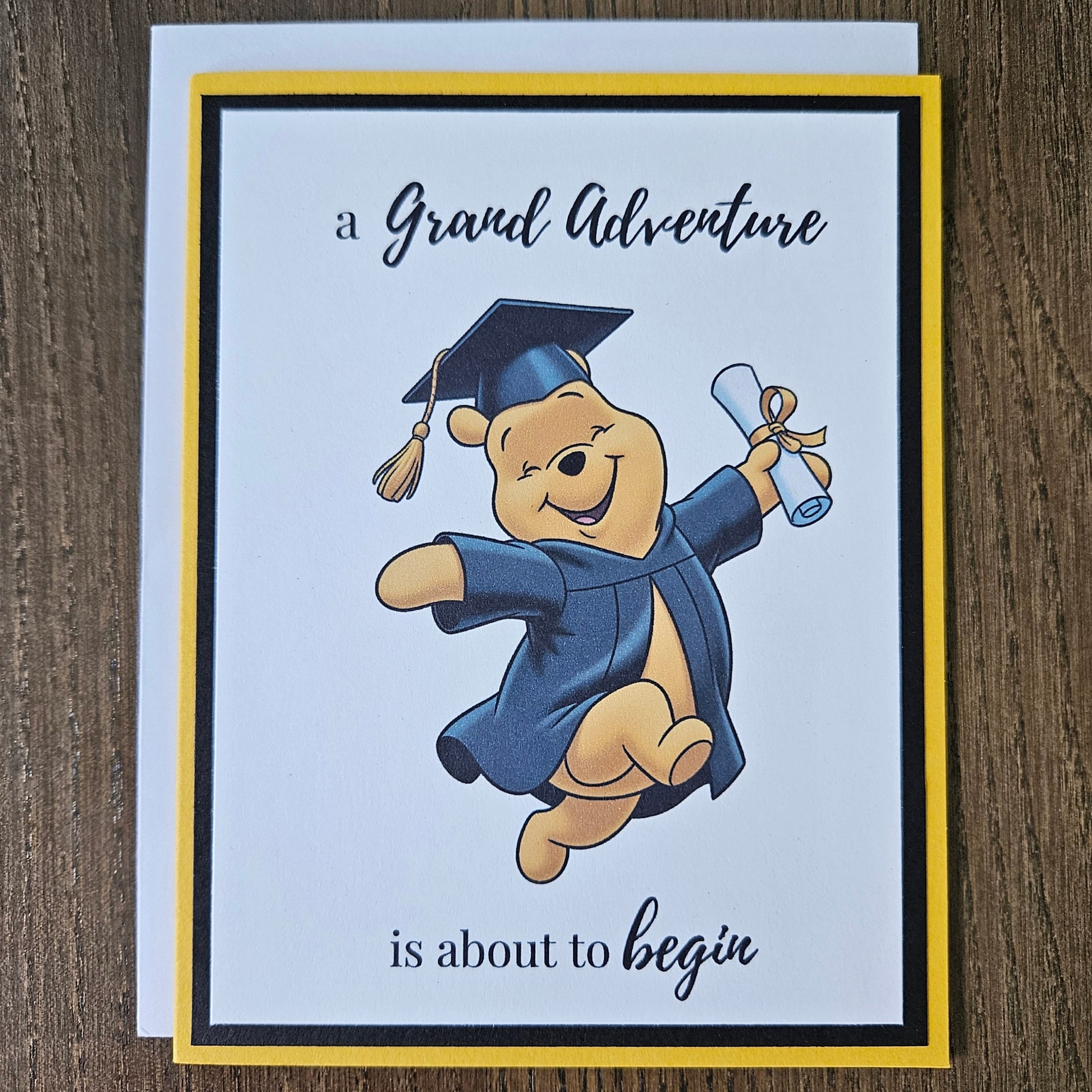 Front of a handmade A2 graduation card featuring a classic storybook sketch illustration of Winnie the Pooh leaping joyfully while wearing a graduation cap and gown and holding a diploma, printed with the text A Grand Adventure is About to Begin, finished with a bold black mat and gold cardstock frame.