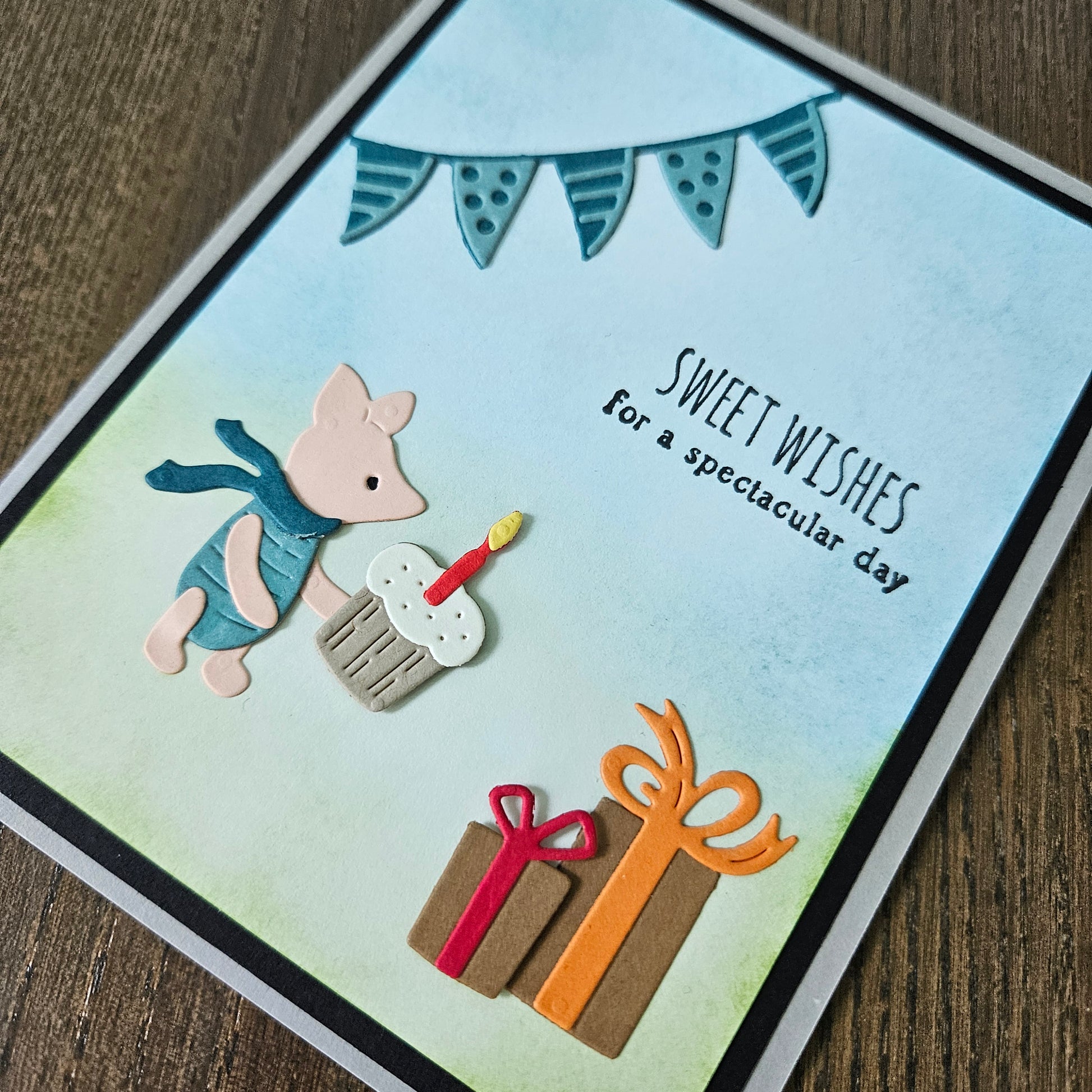 Greeting card with a cartoon pig holding a cake, surrounded by presents and text on a wooden surface.